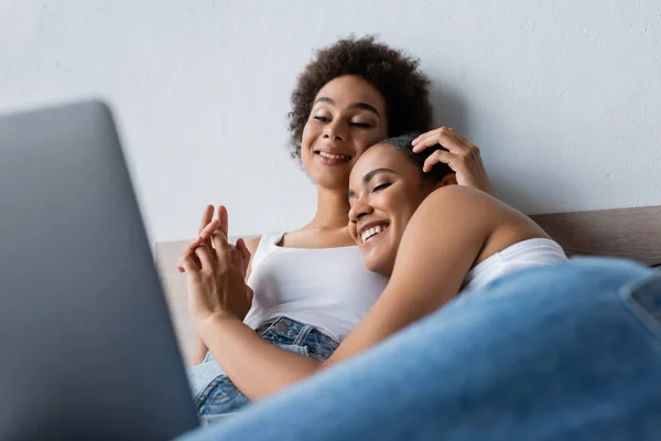 Feliz africano americano lesbiana pareja viendo película en portátil y cogido de la mano en dormitorio - foto de stock
