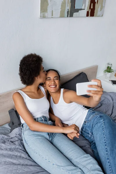 Високий кут зору щасливої афро-американської лесбійської пари, що приймає селфі на смартфон у спальні — стокове фото