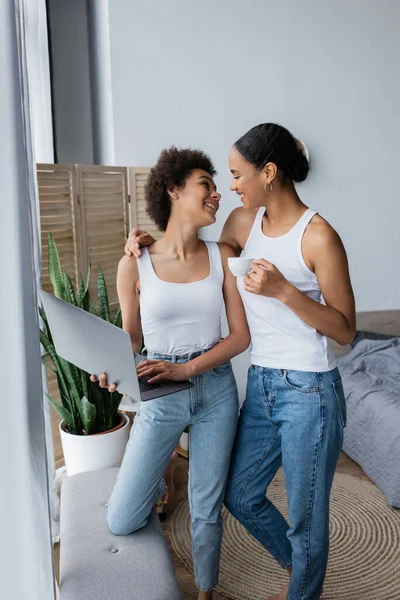 Cheerful african american lesbian woman using laptop while girlfriend holding cup of coffee — Stock Photo