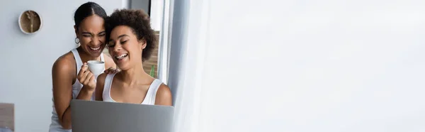 Allegra donna lesbica africana americana che utilizza il computer portatile vicino alla ragazza felice con in mano una tazza di caffè, banner — Foto stock