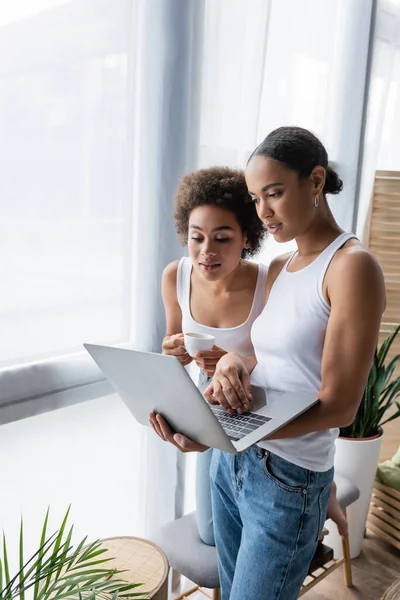 African american lesbian woman using laptop while curly girlfriend holding cup of coffee — Stock Photo