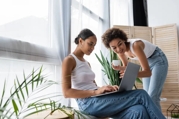 Junge afrikanisch-amerikanische lesbische Frau mit Laptop in der Nähe lockige Freundin hält Tasse Kaffee — Stockfoto