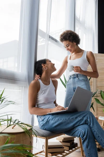Young african american lesbian woman using laptop near smiling girlfriend holding cup of coffee — Stock Photo
