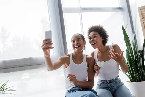 Happy african american lesbian woman taking selfie with girlfriend holding cup of coffee — Stock Photo