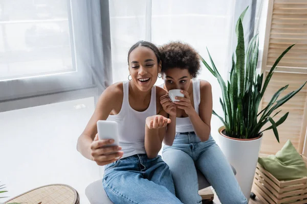 Щаслива афро-американська лесбіянка розмовляє під час відео чату на смартфоні, коли дівчина п'є каву — стокове фото