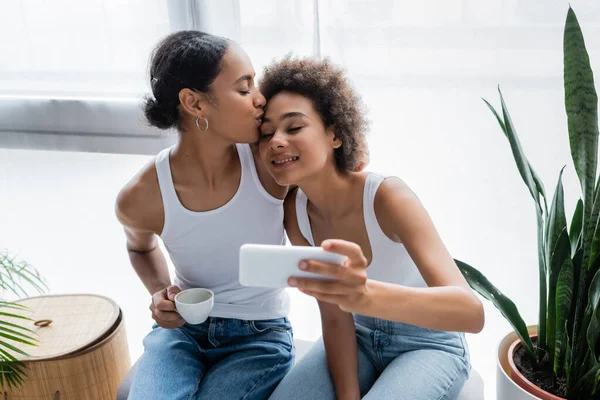 Allegra donna lesbica africana americana che prende selfie mentre la ragazza tiene in mano una tazza di caffè e le bacia la testa — Foto stock