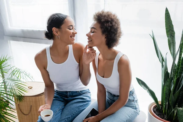 Cheerful african american lesbian woman touching chin of happy partner with smartphone — Stock Photo