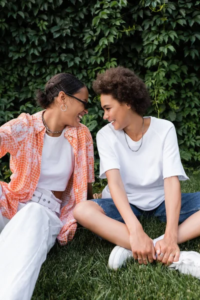 Gai afro-américain lesbienne femme en lunettes de soleil regardant petite amie heureuse tout en étant assis sur la pelouse — Photo de stock