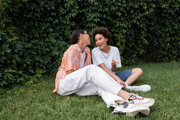 Stylische afrikanisch-amerikanische lesbische Frau mit Sonnenbrille schaut aufgeregte Freundin an, während sie auf dem Rasen sitzt — Stockfoto