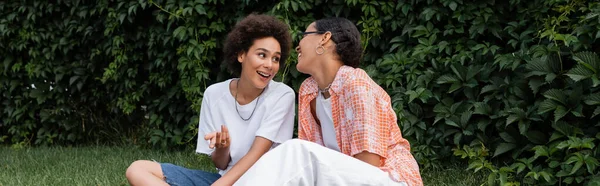 Stylish african american lesbian woman in sunglasses looking at happy girlfriend while sitting on lawn, banner — Stock Photo