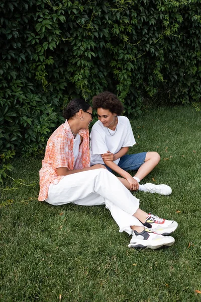Souriant afro-américain lesbienne femme en lunettes de soleil regardant petite amie heureuse tout en étant assis sur la pelouse — Photo de stock