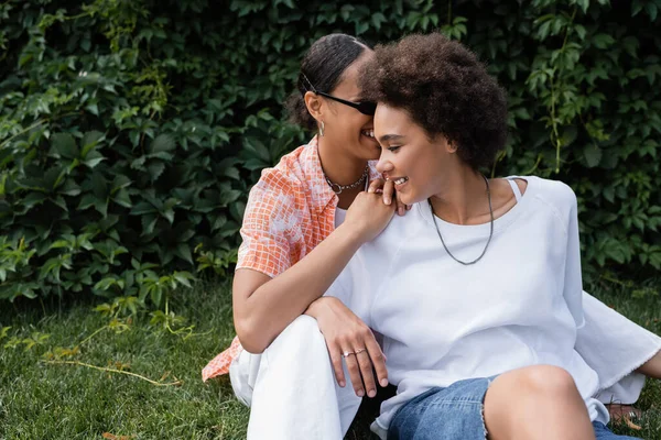 Alegre Africano americano lésbica mulher em óculos de sol abraçando feliz namorada enquanto sentado no gramado — Fotografia de Stock