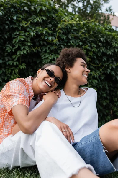 Stylish african american lesbian woman in sunglasses leaning on happy girlfriend while sitting in green park — Stock Photo