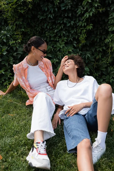 Stylish african american lesbian woman in sunglasses stroking hair of happy girlfriend and sitting on lawn — Stock Photo