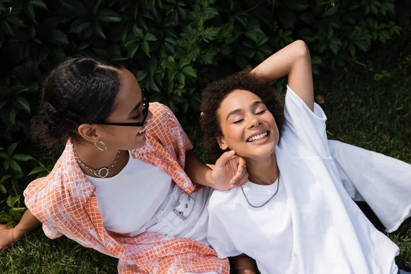 Top view of african american lesbian woman in sunglasses looking at happy girlfriend while sitting on lawn — Stock Photo