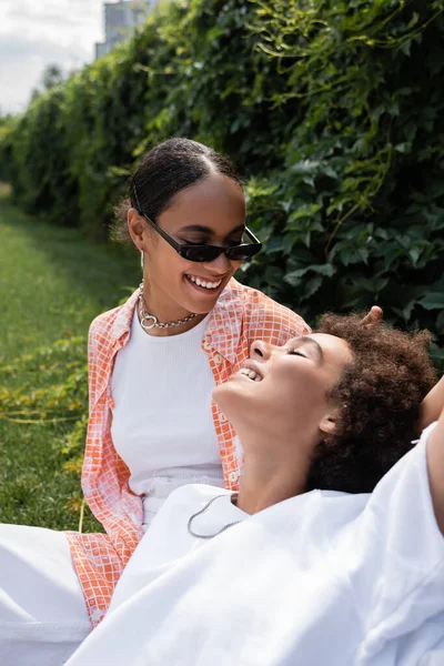 Joyeuse africaine américaine lesbienne femme en lunettes de soleil regardant petite amie heureuse tout en étant assis sur la pelouse — Photo de stock