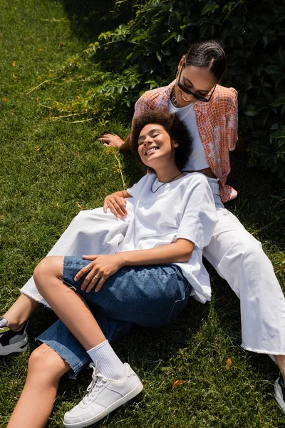 Vue grand angle de femme lesbienne afro-américaine élégante en lunettes de soleil regardant petite amie heureuse dans le parc — Photo de stock