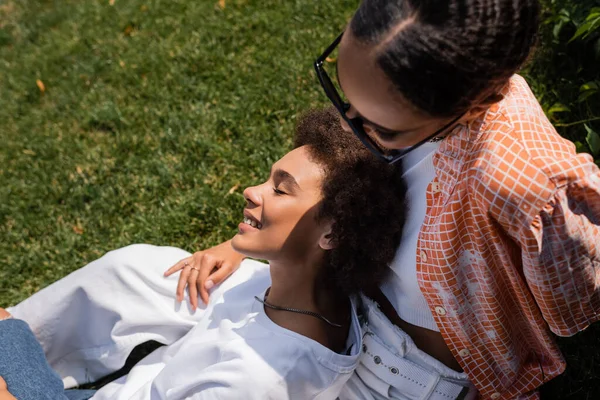 Vista superior de mujer lesbiana afroamericana en gafas de sol abrazando a su novia sonriente mientras está sentada en el césped - foto de stock