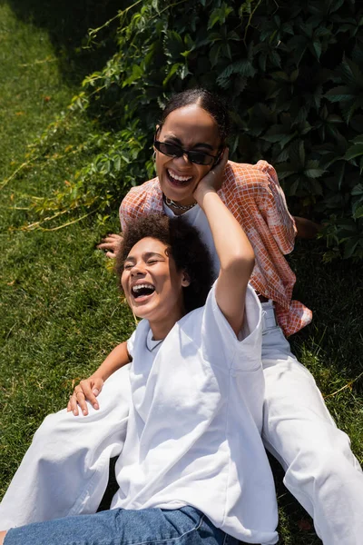 Glücklich afrikanisch-amerikanische lesbische Frau umarmt fröhliche Freundin mit Sonnenbrille, während sie auf dem Rasen sitzt — Stockfoto
