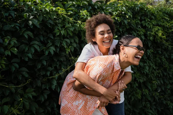 Feliz africana americana lesbiana mujer abrazando excitada novia en gafas de sol riendo en verde parque - foto de stock