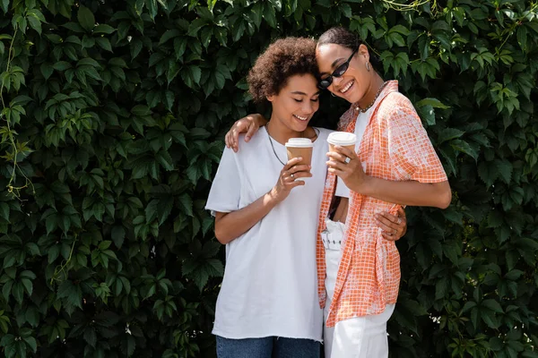 Feliz afroamericana lesbiana mujer en gafas de sol abrazando alegre novia y sosteniendo café para ir - foto de stock