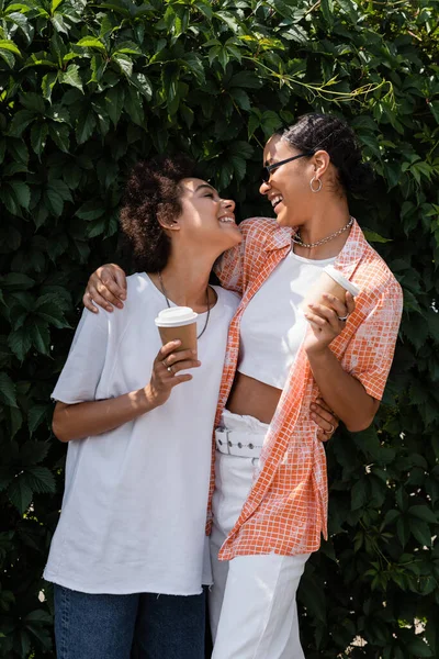 Alegre Africano americano lésbica mulher em óculos de sol abraçando alegre namorada e segurando café para ir — Fotografia de Stock