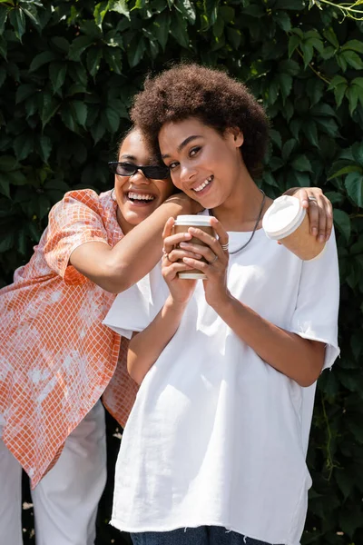 Sonriente afroamericana lesbiana mujer en gafas de sol abrazando rizado novia y sosteniendo café para ir - foto de stock