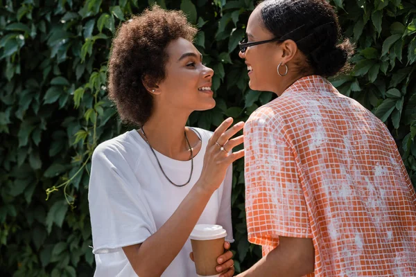 Feliz africana americana lesbiana mujer en gafas de sol mirando rizado novia con café a ir - foto de stock