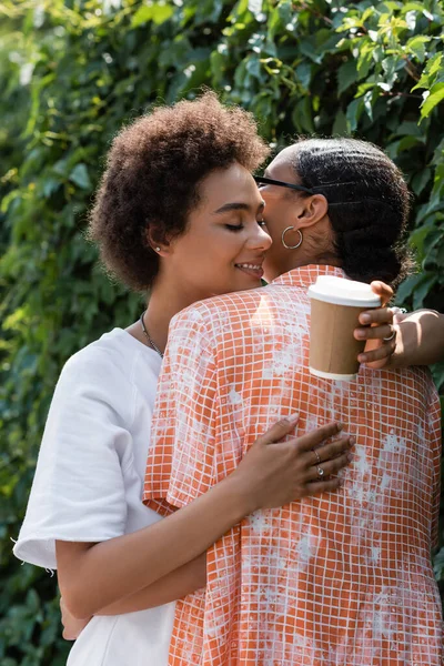 Feliz africana americana lesbiana mujer abrazando novia y sosteniendo café a ir - foto de stock