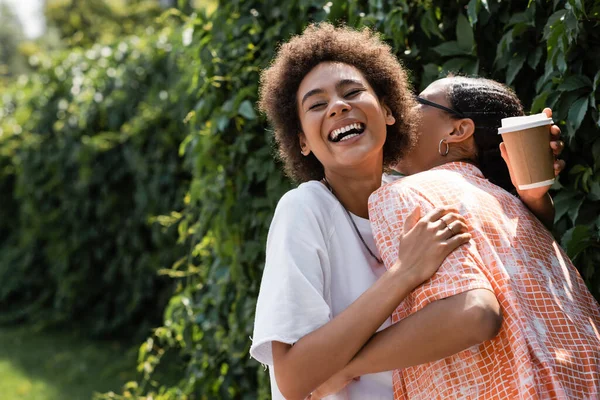 Excitada afroamericana lesbiana mujer abrazando novia y sosteniendo café para ir - foto de stock