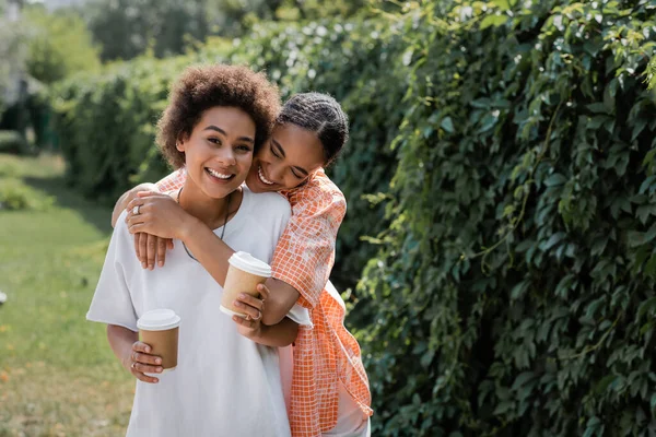 Feliz africano americano lesbiana pareja celebración de papel tazas con café a ir mientras abrazo en parque - foto de stock