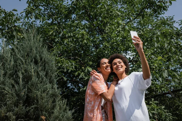 Feliz africano americano lesbiana pareja tomando selfie cerca árbol en verde parque - foto de stock