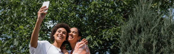 Feliz africano americano lesbiana pareja tomando selfie cerca árbol en verde parque, banner - foto de stock