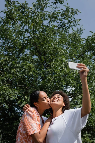 Happy african american lesbian woman kissing cheek of girlfriend while taking selfie near tree in park — Stock Photo