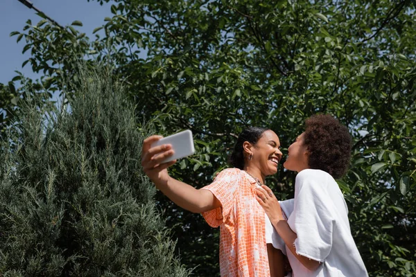 Alegre africano americano lesbiana pareja tomando selfie cerca árbol en verde parque - foto de stock