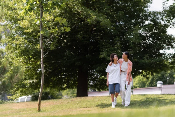 Longitud completa de la joven afroamericana mujer abrazando a su pareja lesbiana y caminando en el parque verde - foto de stock