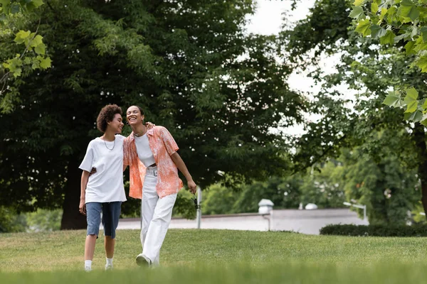Full length of joyful african american lesbian woman hugging happy girlfriend and walking in green park — Stock Photo