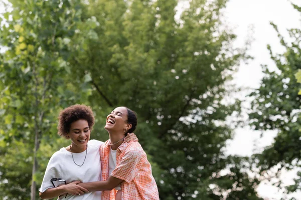 Joyful african american lesbian woman hugging happy girlfriend and laughing in green park — Stock Photo