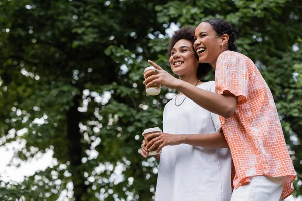 Joyful african american lesbian woman holding coffee to go while pointing away near girlfriend in park — Stock Photo