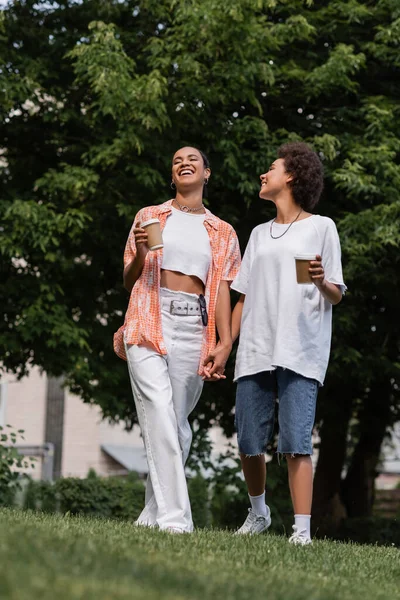 Full length of joyful african american lesbian women holding paper cups and standing in park — Stock Photo
