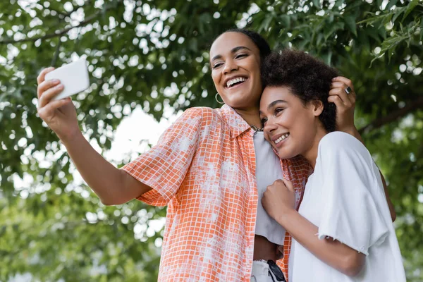 Positivo africano americano lesbiana pareja tomando selfie cerca árbol en verde parque - foto de stock
