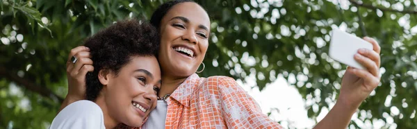 Positivo africano americano lesbiana pareja tomando selfie cerca árbol en verde parque, banner - foto de stock
