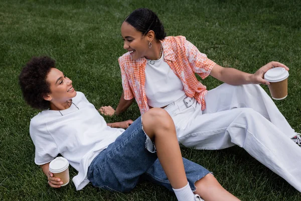 Joyful african american lesbian women holding paper cups and resting on grass in park — Stock Photo