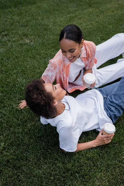 Vista aérea de alegres mujeres lesbianas afroamericanas sosteniendo vasos de papel y sentadas en la hierba en el parque - foto de stock