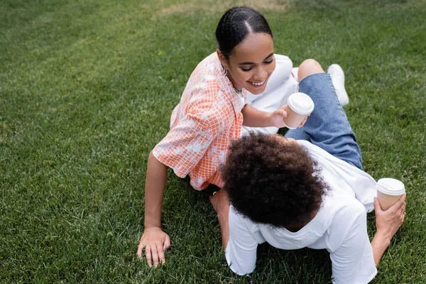 Joyful african american lesbian woman holding paper cup and looking at curly girlfriend sitting on grass in park — Stock Photo