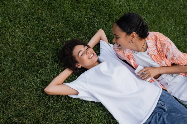 Draufsicht auf freudige afrikanisch-amerikanische lesbische Frau mit Smartphone und Blick auf Freundin im Gras liegend — Stockfoto