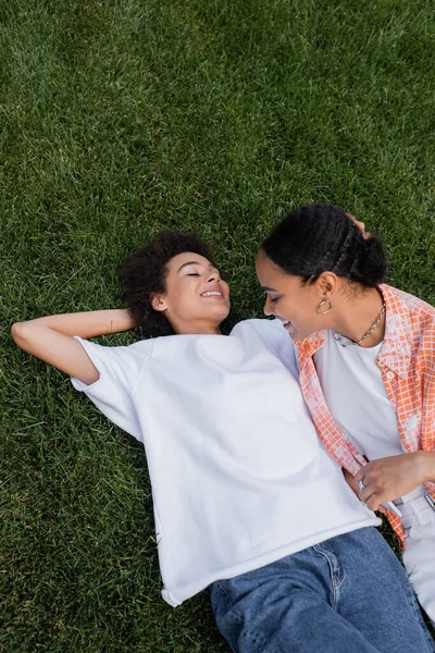 Вид сверху на счастливую американскую лесбиянку из Африки, держащую смартфон рядом с девушкой, лежащей на траве — стоковое фото