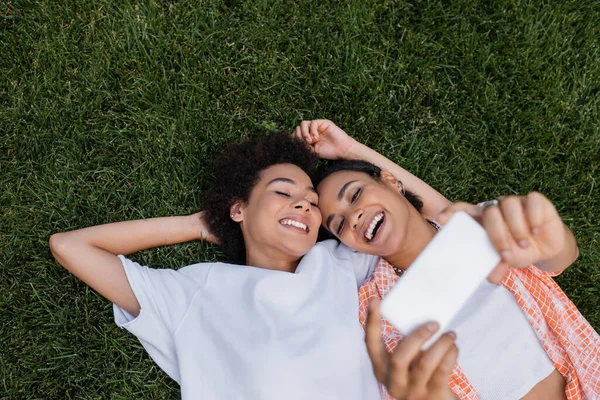 Vista superior de la alegre mujer lesbiana afroamericana sosteniendo el teléfono inteligente y tomando selfie con la novia acostada en la hierba - foto de stock