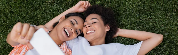 Vue du haut de joyeuse femme lesbienne afro-américaine tenant smartphone et prenant selfie avec sa petite amie allongée sur l'herbe, bannière — Photo de stock
