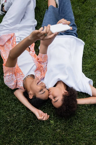 Вид сверху на счастливую американскую лесбиянку из Африки, держащую смартфон и делающую селфи с подружкой, лежащей на траве — стоковое фото
